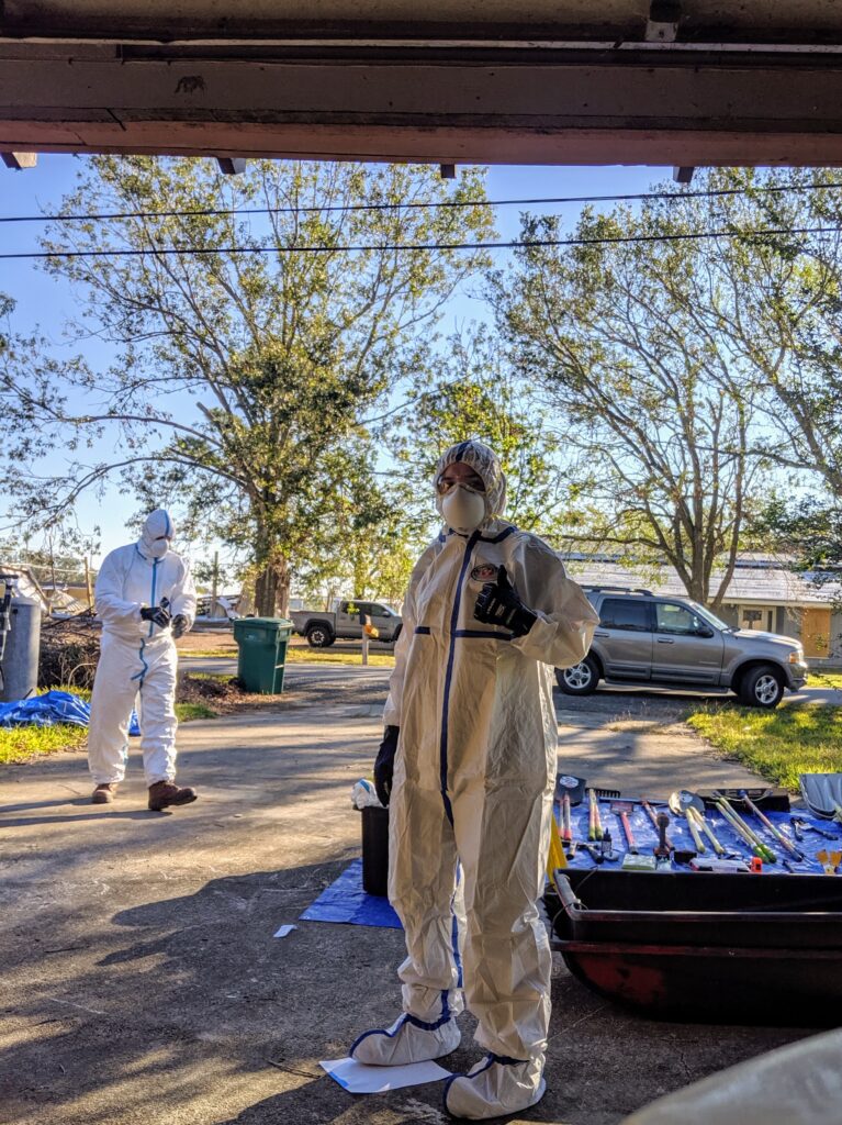 What appears to be a human covered from head to toe in Tyvek and PPE stands in a driveway with an assortment of demolition tools behind it.